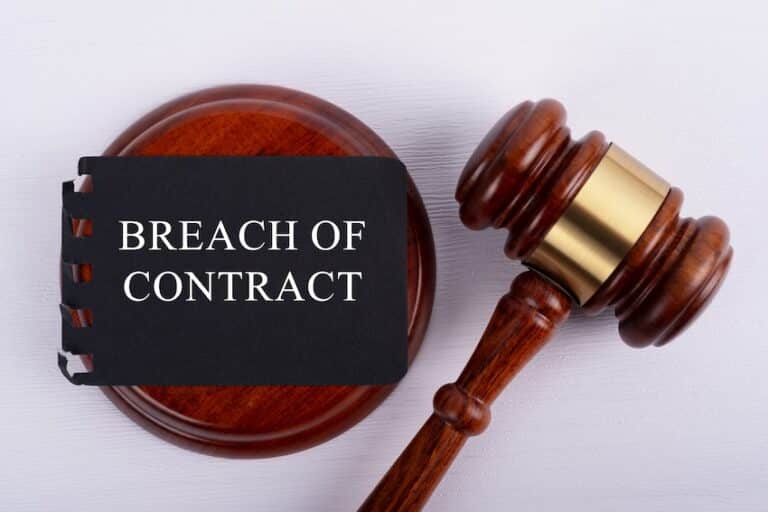 A small black spiral-bound notepad with torn and slightly crumpled edges sits propped against a round polished mahogany judge's sound block, printed with the words "BREACH OF CONTRACT" in bold white capital letters across its dark cover while a warm honey-toned wooden gavel with a shiny gold band rests diagonally beside it on a clean white textured surface — a straightforward legal concept image representing the serious consequences of a material breach in a contractual agreement and could use help from Rosenbaum & Taylor P.C.