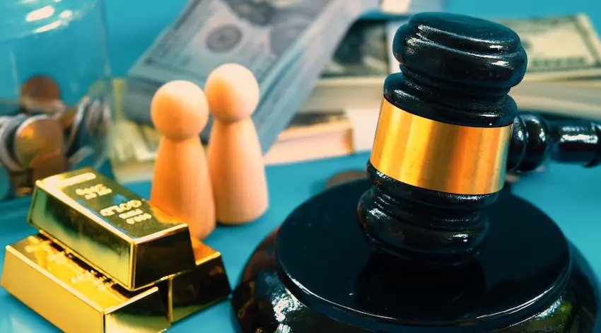 A black judge's gavel with a gold band rests on its circular black sound block on a vivid teal surface positioned beside two small rounded wooden peg figurines standing upright to represent opposing parties- two stacked gold bullion bars etched with fine detail- a loose pile of mixed coins partially contained in a clear glass vessel- and a thick stack of bundled hundred-dollar bills in the blurred background — a conceptual arrangement illustrating the significant financial stakes and legal consequences involved in a material breach dispute in New York.