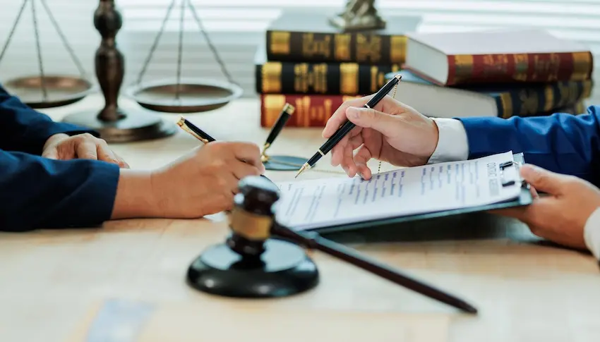 Two people seated across from each other at a light wood conference table both hold black and gold ballpoint pens over a document on a clipboard with one person in a blue jacket pointing to a specific line on the page while the other prepares to sign- a small black wooden judge's gavel lying flat in the foreground- a brass balance scale of justice and a tall stack of dark law books with gold lettering visible in the softly blurred background — depicting the formal legal review and documentation process that follows a material breach of contract in New York.
